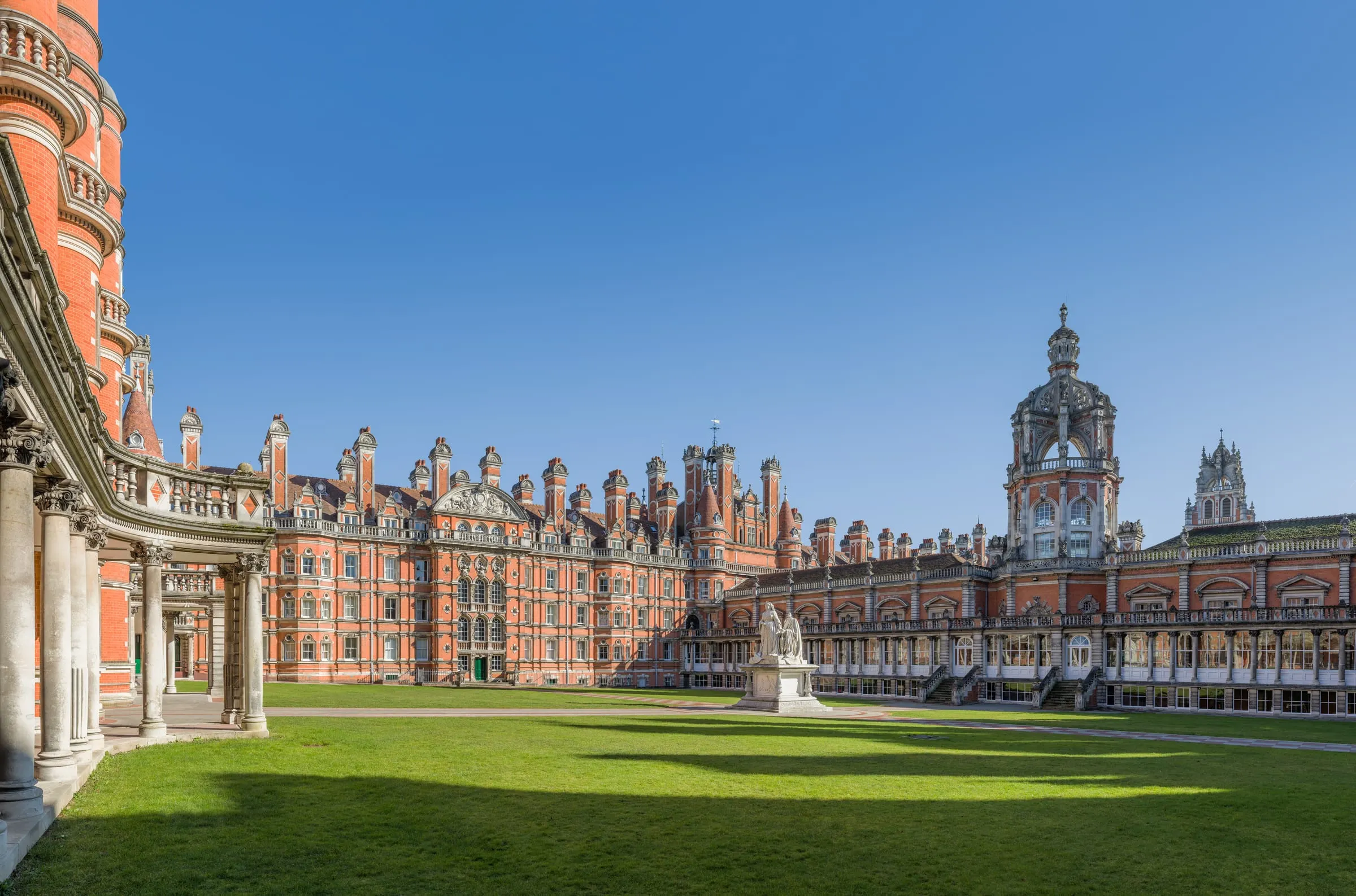 Edificio victoriano de ladrillo rojo con torres y patio de césped, sede de un campus universitario cerca de Londres