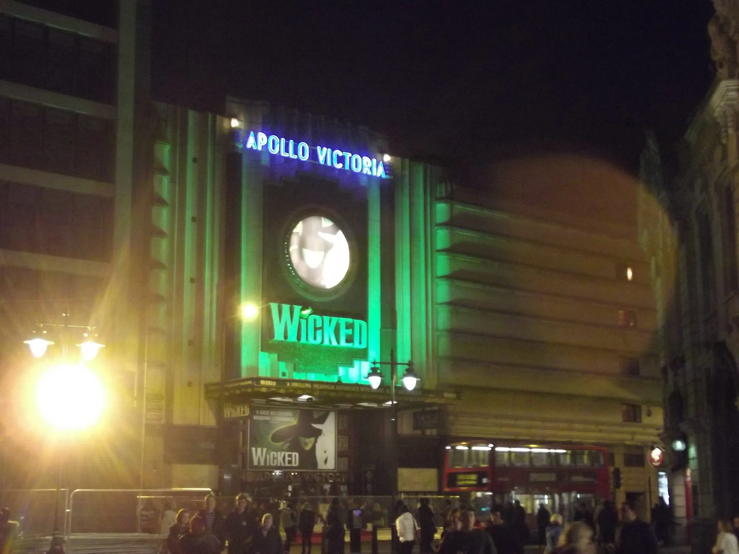 El Apollo Victoria iluminado de verde por la noche, con el cartel de Wicked y un autobús rojo